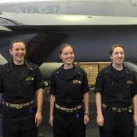 Three women in black jumpsuits stand in front of an aircraft below the decks of an aircraft carrier