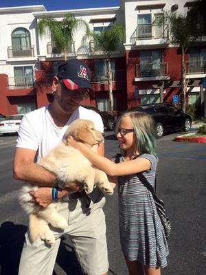 A man wearing a UA baseball cap holds a small fluffy labrador puppy, while a young girl with blue stripes in her hair pets the dog's face.