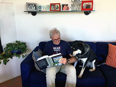 A man in a blue UA shirt sits on a blue couch next to a large black and white dog. He appears to be reading aloud to the dog from a book.