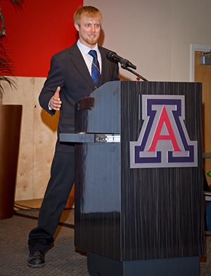 A man in a dark business suit and a blue tie stands at a podium with the University of Arizona logo on it