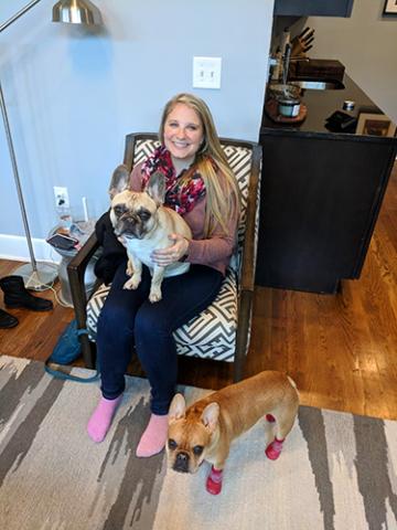 A woman sitting in a chair and holding a French bulldog while another French bulldog stands by her feet