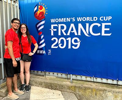 Christian Montoya and Jessyca Thomas wearing red shirts and standing in front of a blue sign that reads "Women's World Cup: France 2019"
