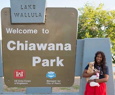 A woman wearing a red skirt and blue UA shirt holding a small yellow dog stands in front of a "Welcome to Chiawana Park" sign