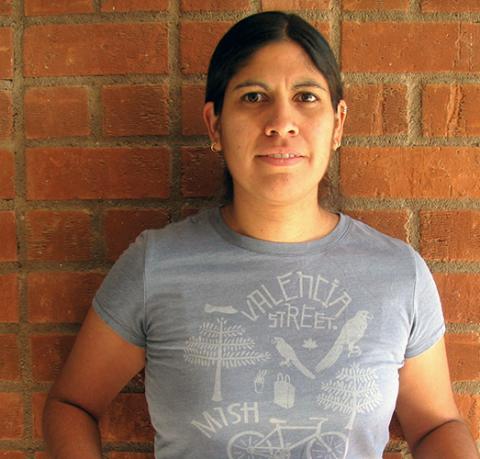 A woman wearing a gray T-shirt and standing before a brick wall.