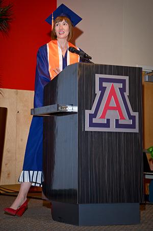 A woman standing at a podium in a blue graduation cap and gown with an orange stole