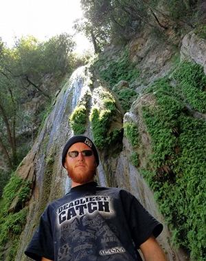 A man with a bushy red beard wearing a black hat stands in front of a waterfall