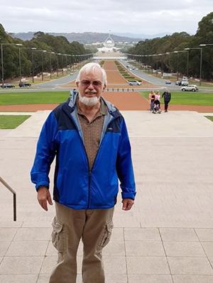 A man with a bushy white beard wearing a blue jacket stands in front of an expansive promenade