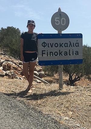 A woman wearing a baseball cap stands in front of a road sign with Greek and English text