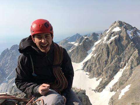 A man in climbing gear, laughing as he sits at the summit of a mountain and other mountains are visible in the background