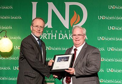 A smaller version of the photo above, wherein a man accepts a framed certificate from another man in front of a University of North Dakota backdrop.
