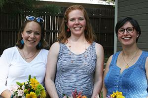Three young smiling women hold bright yellow flowers in front of their pregnant stomachs.