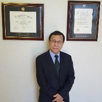 A man in a black suit and wire-rimmed glasses stands in front of two framed diplomas