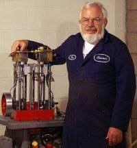 A man with a white beard wearing a shop jumpsuit stands in front of a handbuilt boiler