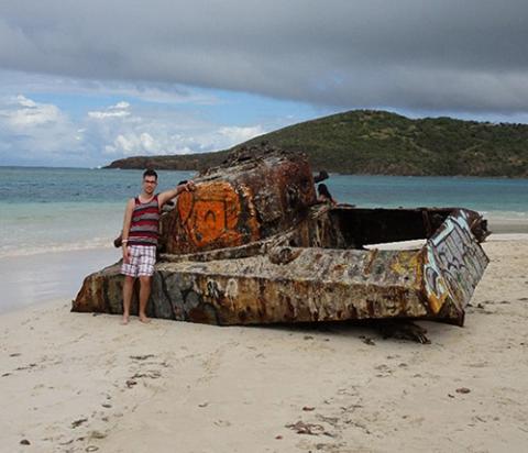 A man in a swim shirt and board shorts stands with a rusted, painted tank on a beach.