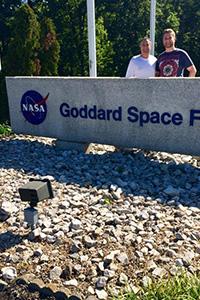 A younger man stands with his father behind a Goddard Space Flight Center entrance sign with the NASA logo