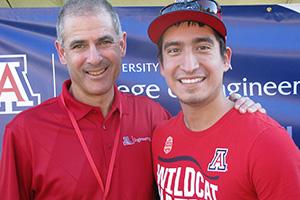 A closer crop of the top photo, in which two mean wearing red UA shirts smile in front of a College of Engineering banner