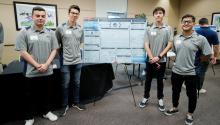 Four students in matching gray polos stand on either side of their research project poster