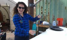 Vicky Karanikola pouring water into a water bottle and smiling. She is outside, wearing sunglasses, and standing in front of a tent and a green metal wall.