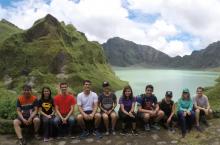 A group of student sitting before a lake that is surrounded by lush green hills.