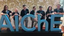 Seven students in blue polo shirts stand behind cut-out letters spelling "AIChE"