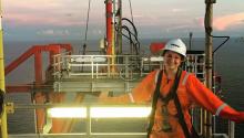 A woman in a bright orange jumpsuit and hard hat stands on the deck of a ship at sea
