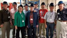 Eight men and one woman stand a laboratory with pipes visible behind them. They are wearing hard hats and lanyards.