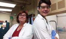 A woman and a young man in lab coats and protective eyewear stand in a chemistry lab with their backs against one another.