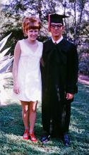 An old photo of a woman with red hair wearing a white dress and red shows standing with a man in a graduation cap and gown