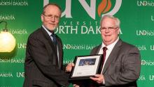 A man accepts a framed certificate from another man in front of a green University of North Dakota backdrop