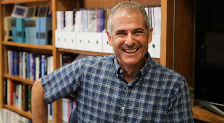 A man in a plaid shirt smiles while sitting in front of a bookshelf.