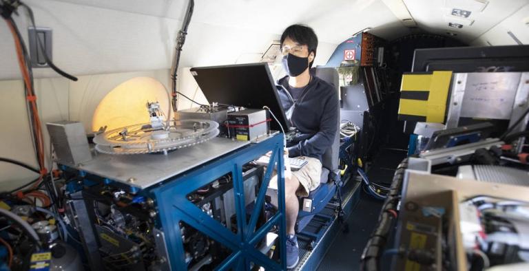A young man wearing a face masks sits behind a monitor on an airplane, surrounded by technology.