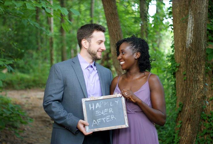 A man and a woman standing in a forest, looking at each other and smiling while they hold a small chalk tablet that reads "happily ever after"