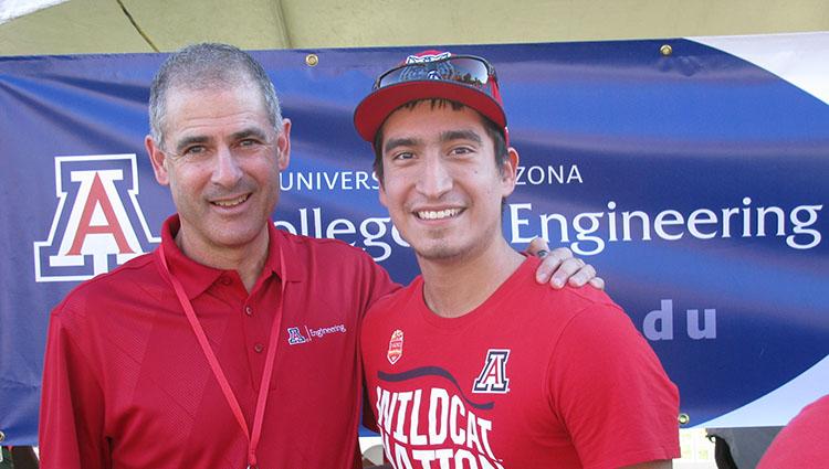 Two men in red University of Arizona shirts smile in front of a blue College of Engineering banner