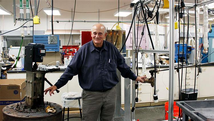 Don Gervasio, wearing a long-sleeved blue shirt, stands between a pump and pipe in a laboratory with tubes hanging in the background.