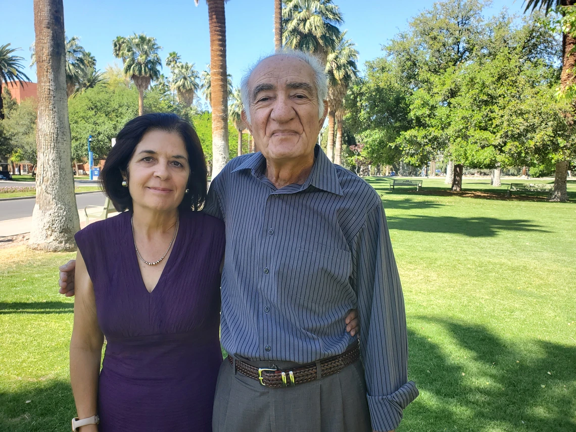 two people, standing and smiling, palm trees and grass in background