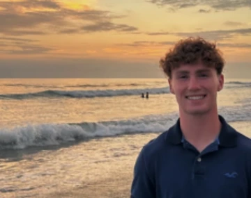 person with short, curly hair wearing blue shirt, ocean at sunset in background