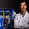 A man with dark hair in a lab photo stands i front of a blue machine.